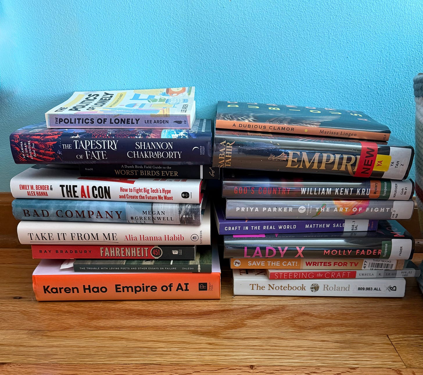 Two stacks of books on a hardwood floor in front of a bright blue wall, with titles including The Politics of Lonely by Lee Arden and A Dubious Clamor by Marissa Lingen Two stacks of books on a hardwood floor in front of a bright blue wall, with titles including The Politics of Lonely by Lee Arden and A Dubious Clamor by Marissa Lingen