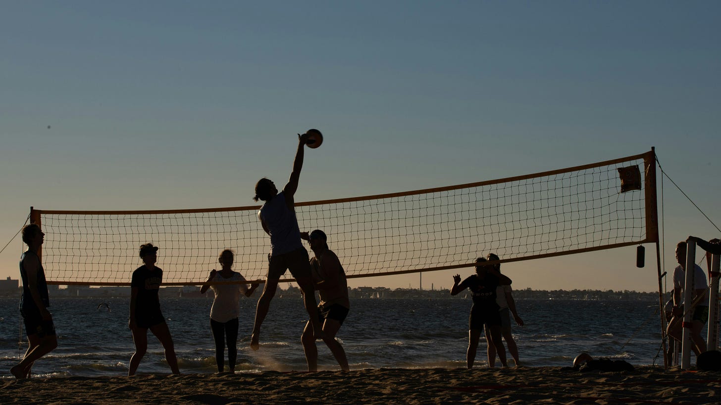 At sunset a group of people (in silhouette) play volleyball on the beach. One person has leaped into the air to spike the ball over the night.