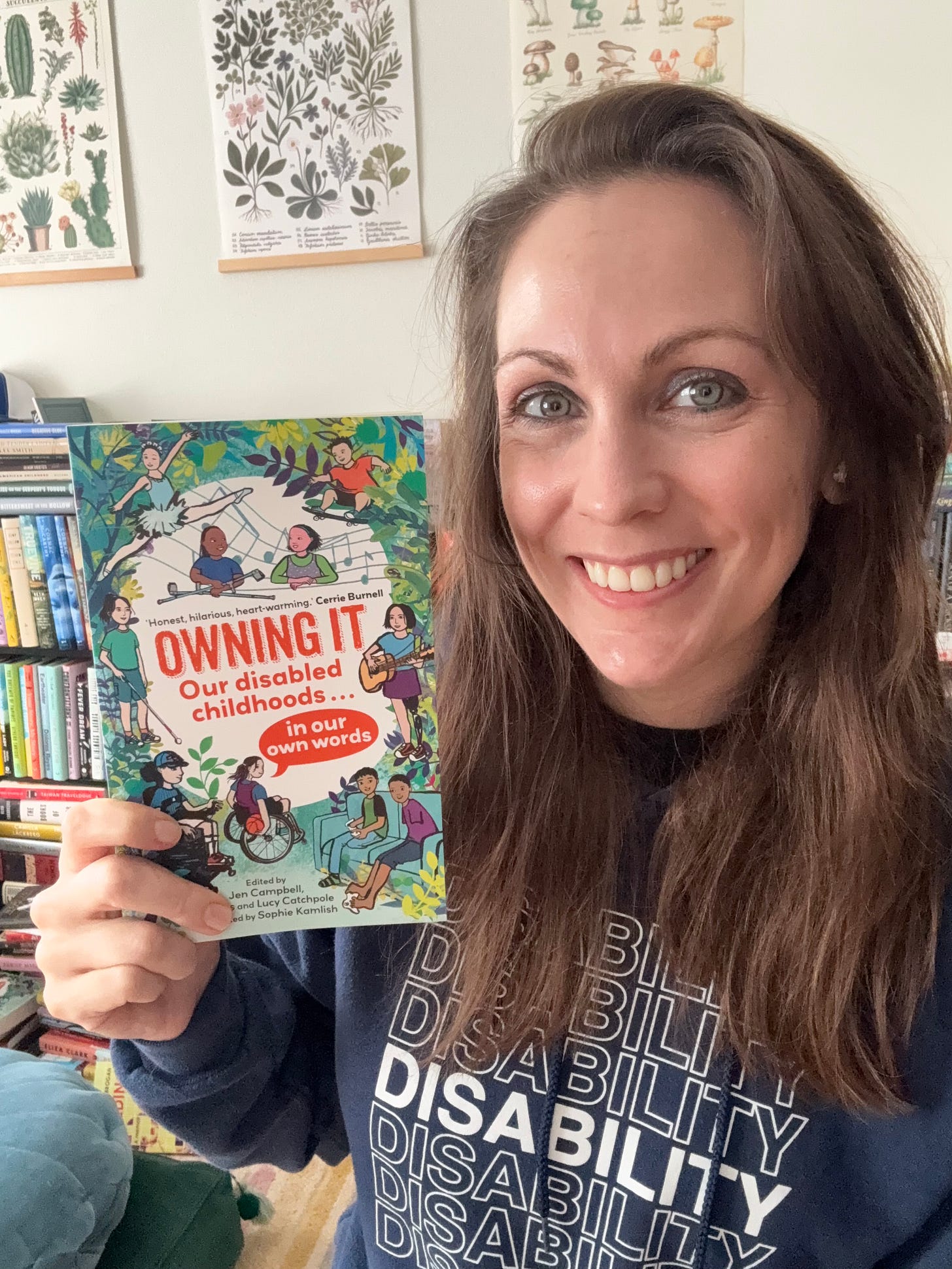 a photo of Kendra, a white thirty-something with long brunette hair, holding a copy of Owning It. She’s wearing a disability sweatshirt and rows of bookshelves can be seen behind her. a photo of Kendra, a white thirty-something with long brunette hair, holding a copy of Owning It. She’s wearing a disability sweatshirt and rows of bookshelves can be seen behind her.