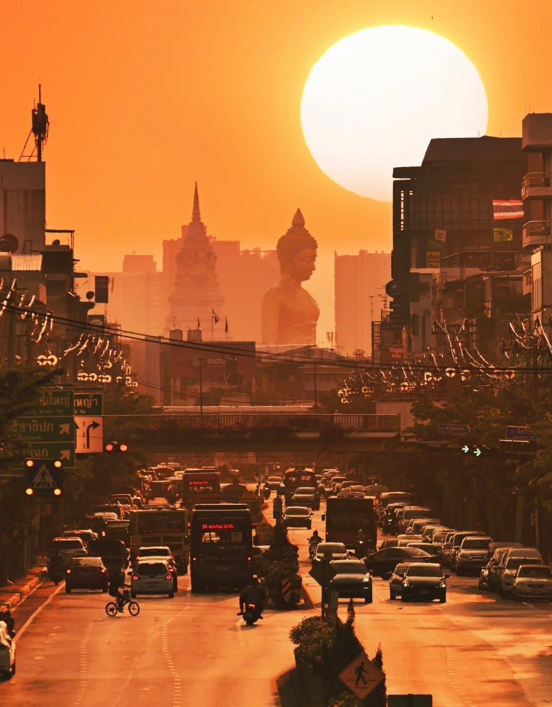 A giant golden Buddha statue and city buildings silhouetted against a large orange sunset over Bangkok, above a busy street full of cars, buses, and motorbikes.