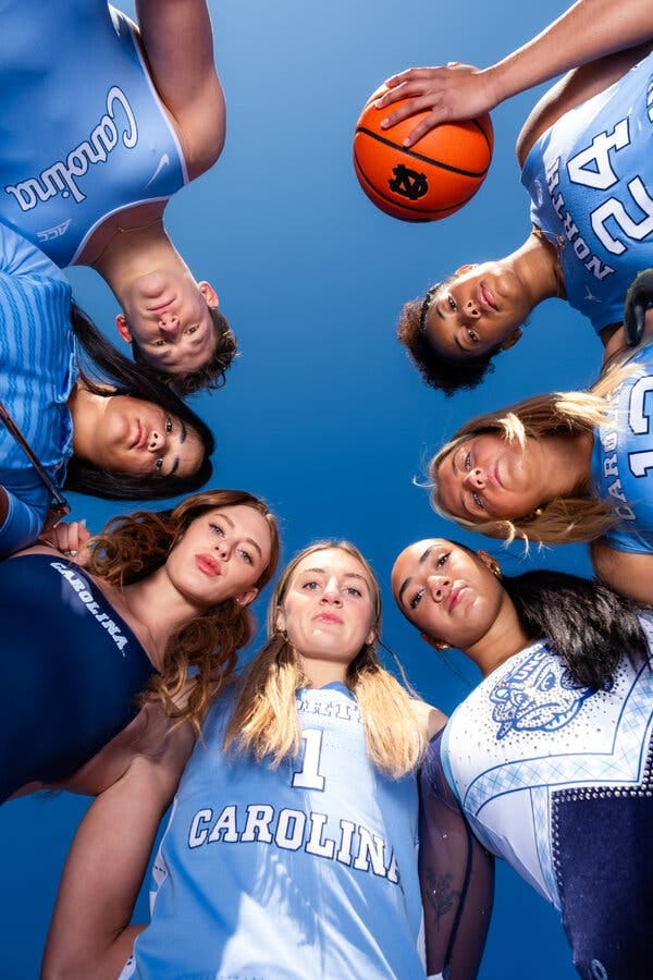 Seven athletes wearing blue U.N.C. gear are huddled in a circle and looking down at the camera. One is holding a U.N.C.-branded basketball.