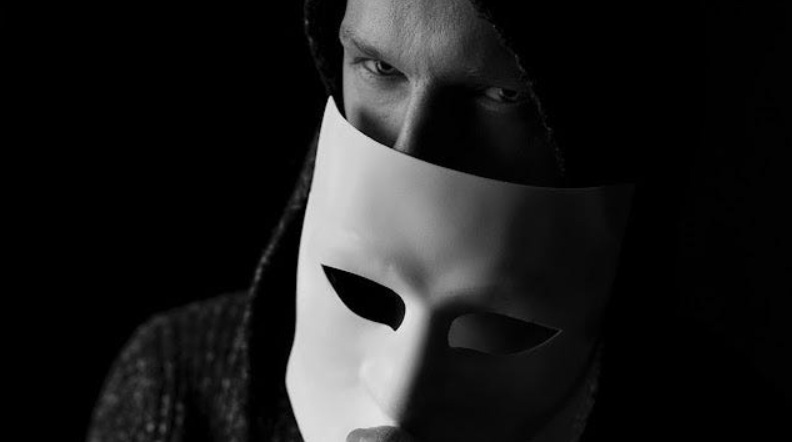 A black and white image displays a person’s eyes peering intensely from a dark hood while holding a stark, white theatrical mask over the lower half of their face. A black and white image displays a person’s eyes peering intensely from a dark hood while holding a stark, white theatrical mask over the lower half of their face.