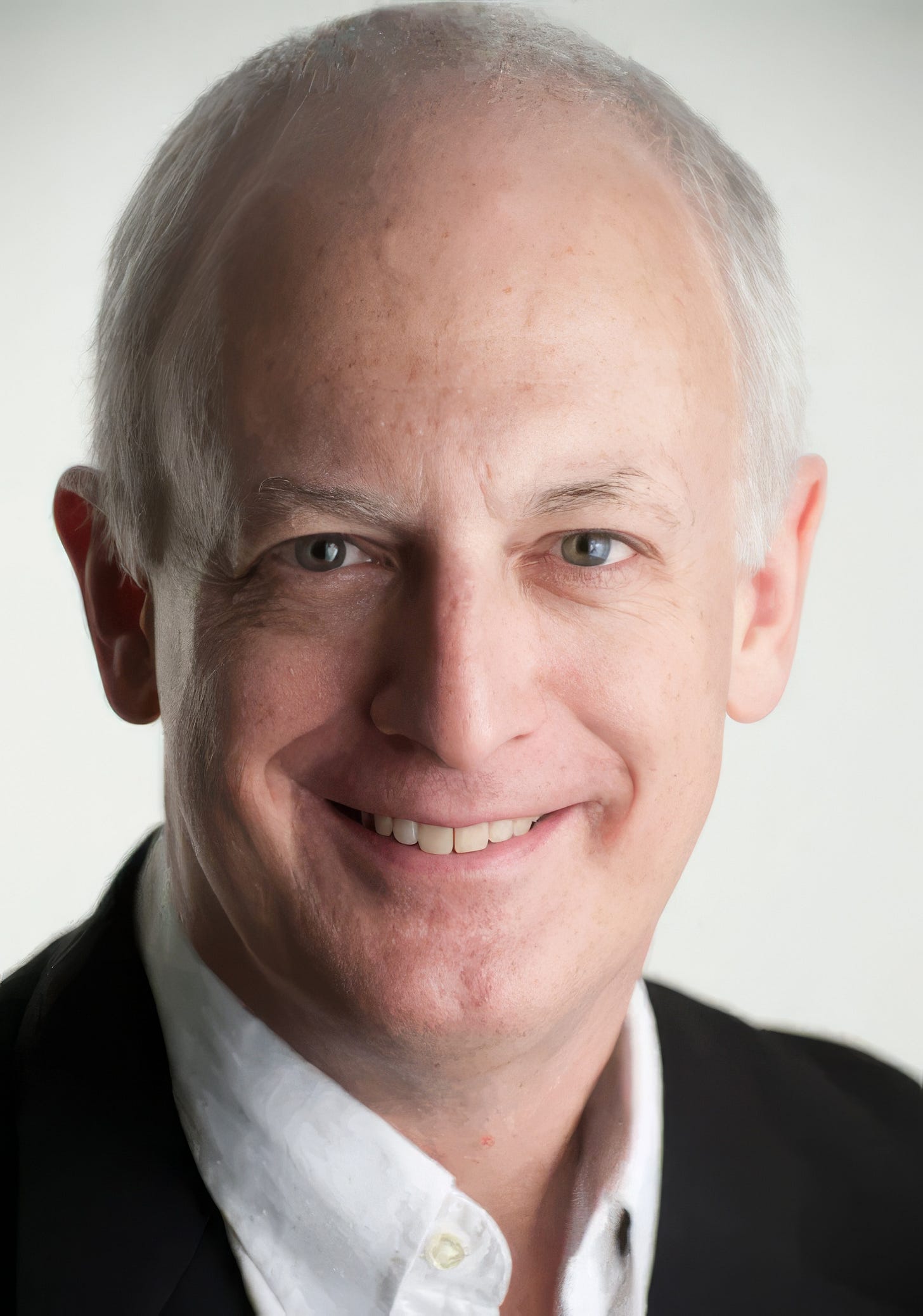 Professional head-and-shoulders portrait of a smiling older man with short grey hair, wearing a black jacket over a white shirt against a light grey background. Professional head-and-shoulders portrait of a smiling older man with short grey hair, wearing a black jacket over a white shirt against a light grey background.