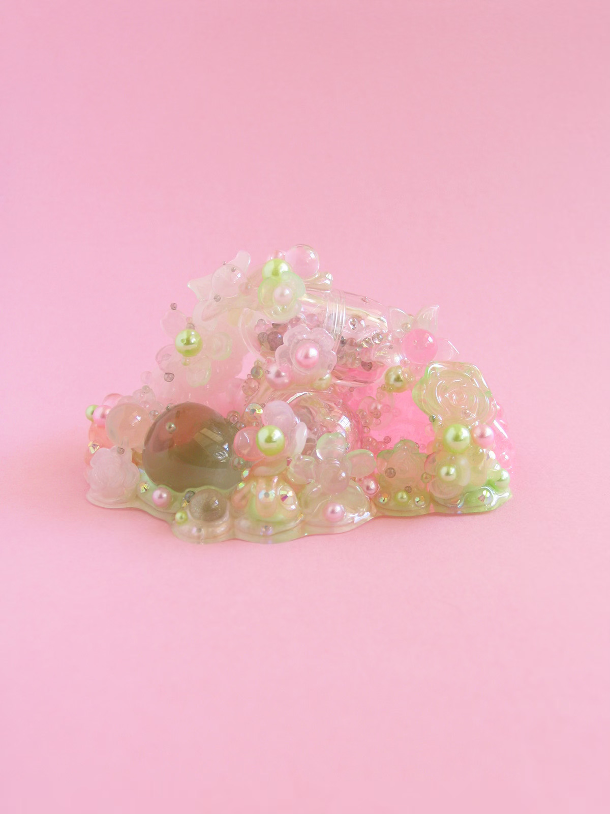 A miniature Glue Baby sculpture: pink and green flowers and crystals melting together on a pink background