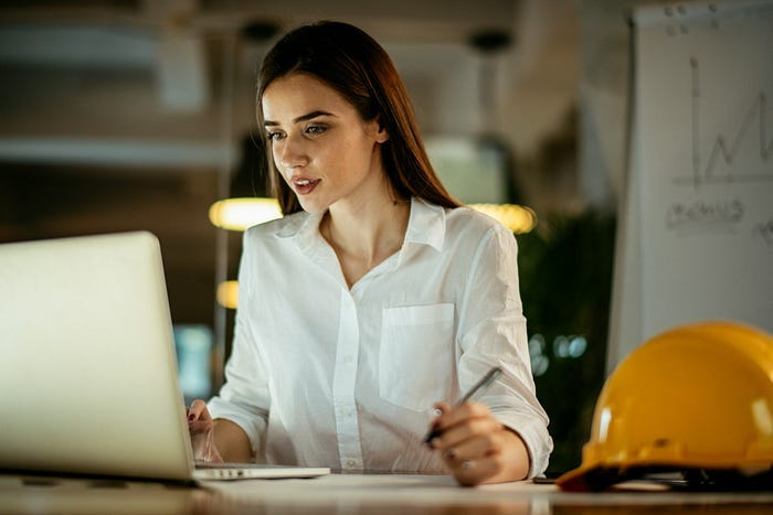 A woman working at a computer next to a hard hat. A woman working at a computer next to a hard hat.
