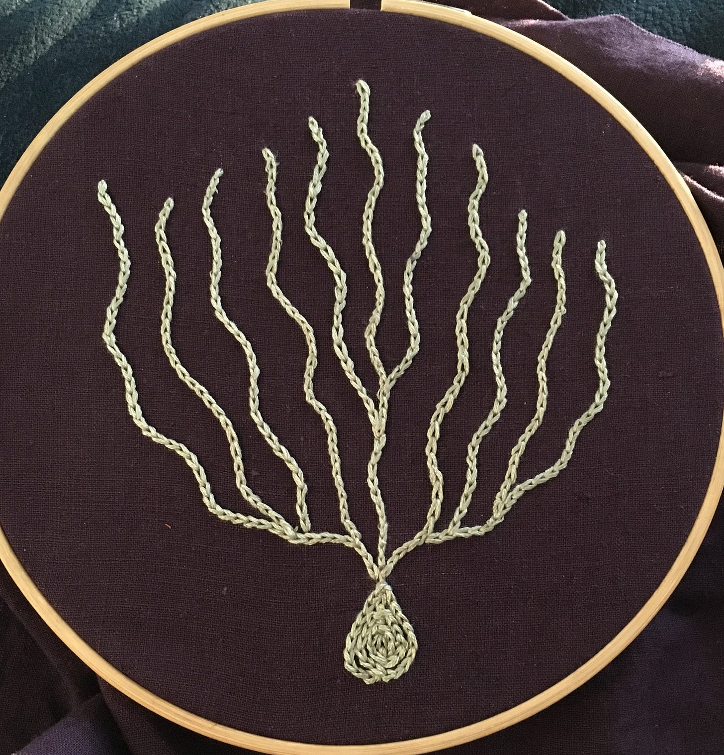 A piece of purple linen in an embroidery hoop. Stitched in chain stitch is a light greenish blue coral or plant or algae design.