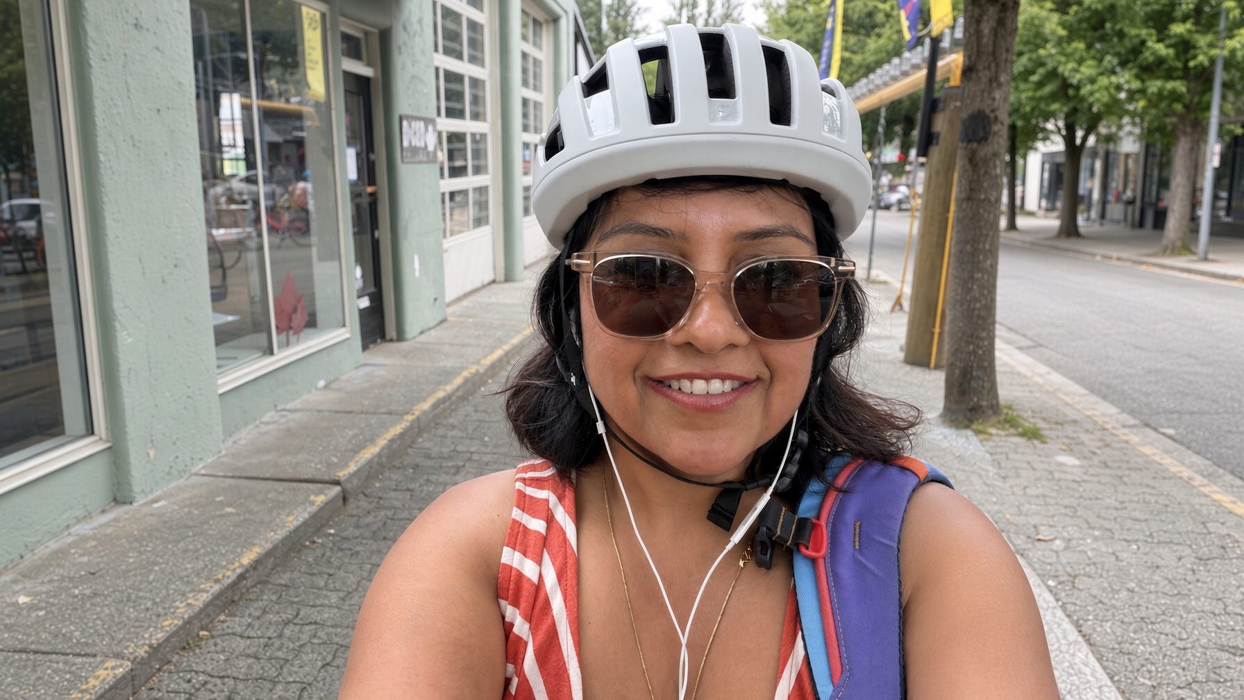 Deepa smiling in a bike helmet and sunglasses on a tree-lined Vancouver street, earbuds in, mid-ride