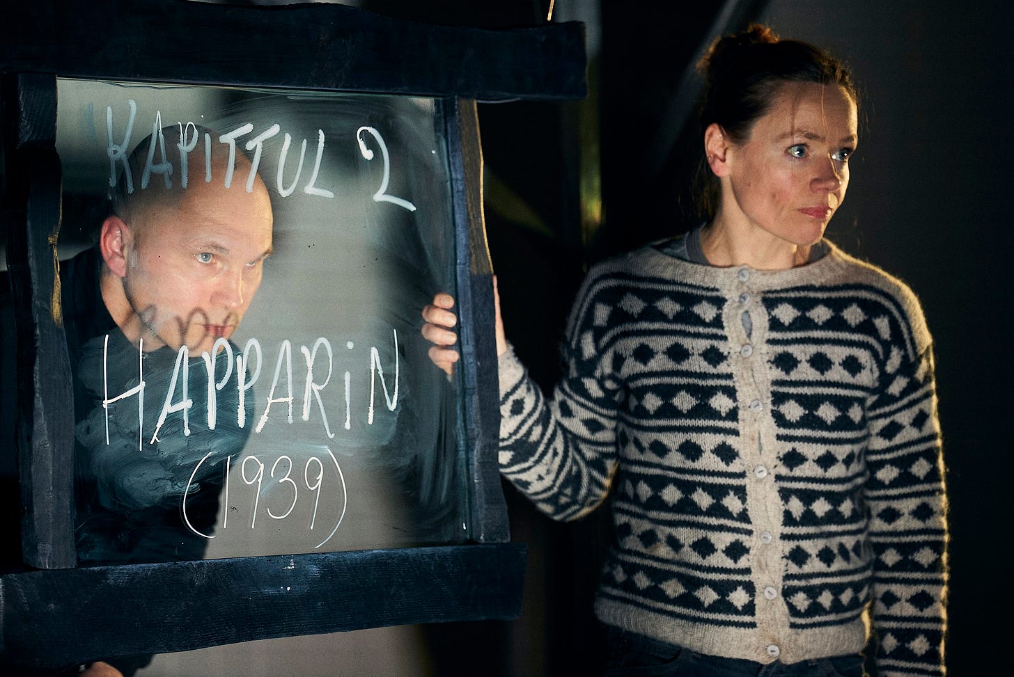 A woman in a cardigan stands next to a window. A man peers through.