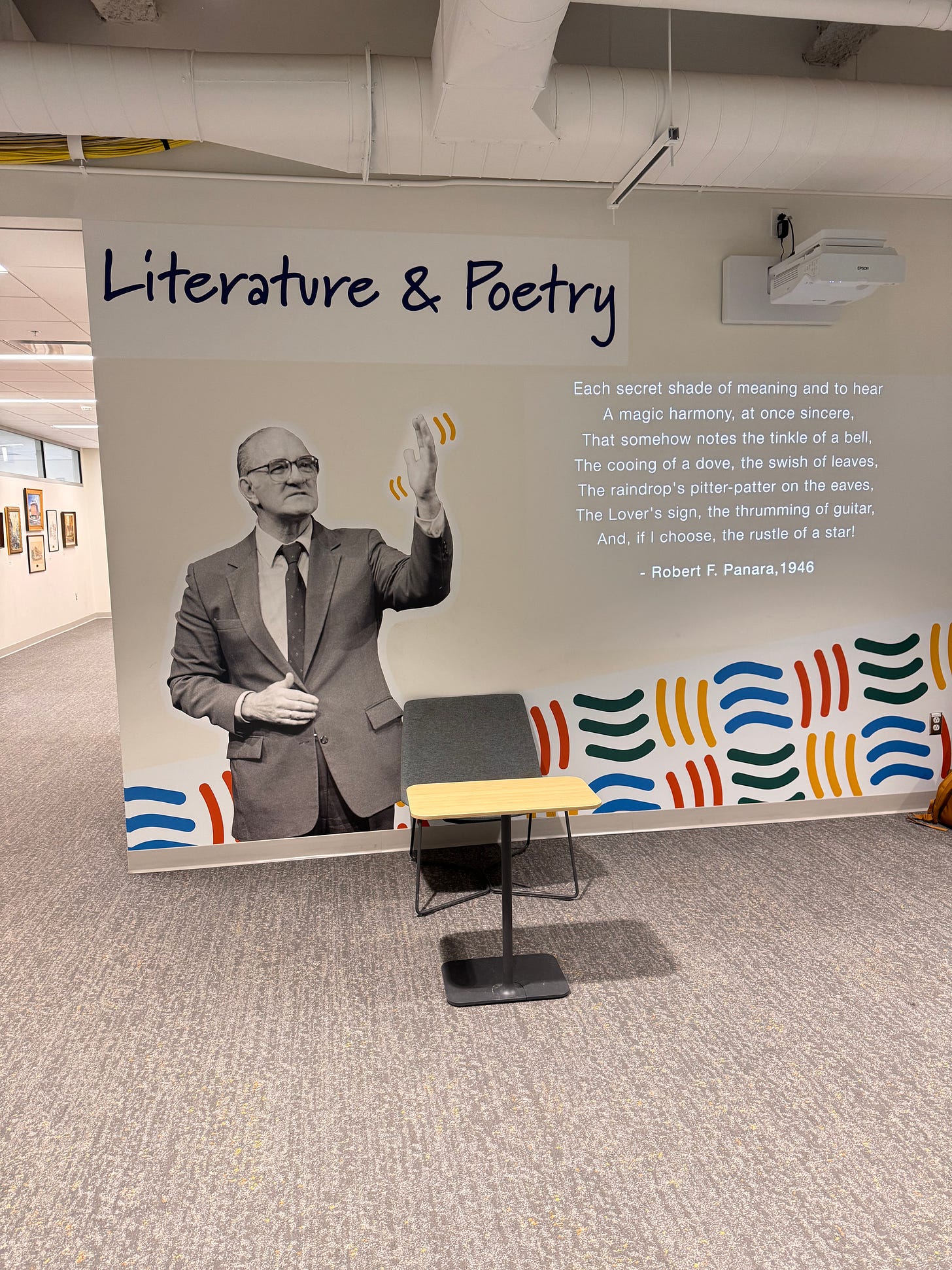Photo from the Robert Panara exhibit at RIT. A black-and-white photo of Panara in a suit is against a white wall, with the header “Literature and Poetry” above his head. To the side are several written lines from Panara’s poem “On His Deafness.”