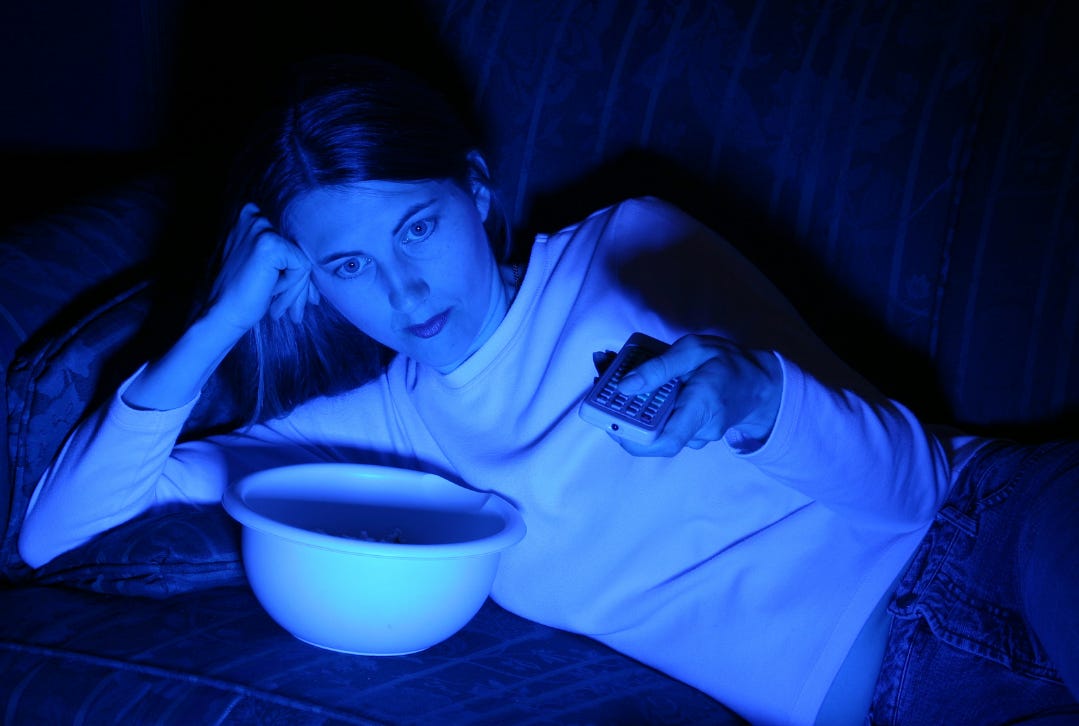 A woman lies on a couch late at night, remote in hand, illuminated by the glow of a television, an empty bowl in front of her. A woman lies on a couch late at night, remote in hand, illuminated by the glow of a television, an empty bowl in front of her.