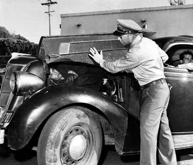 A man with a police hat lifts up the hood of an old car to reveal a man lying there in a black and white photo. A man with a police hat lifts up the hood of an old car to reveal a man lying there in a black and white photo.