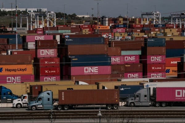 A line of trucks in the foreground. Behind them are piles of shipping containers.