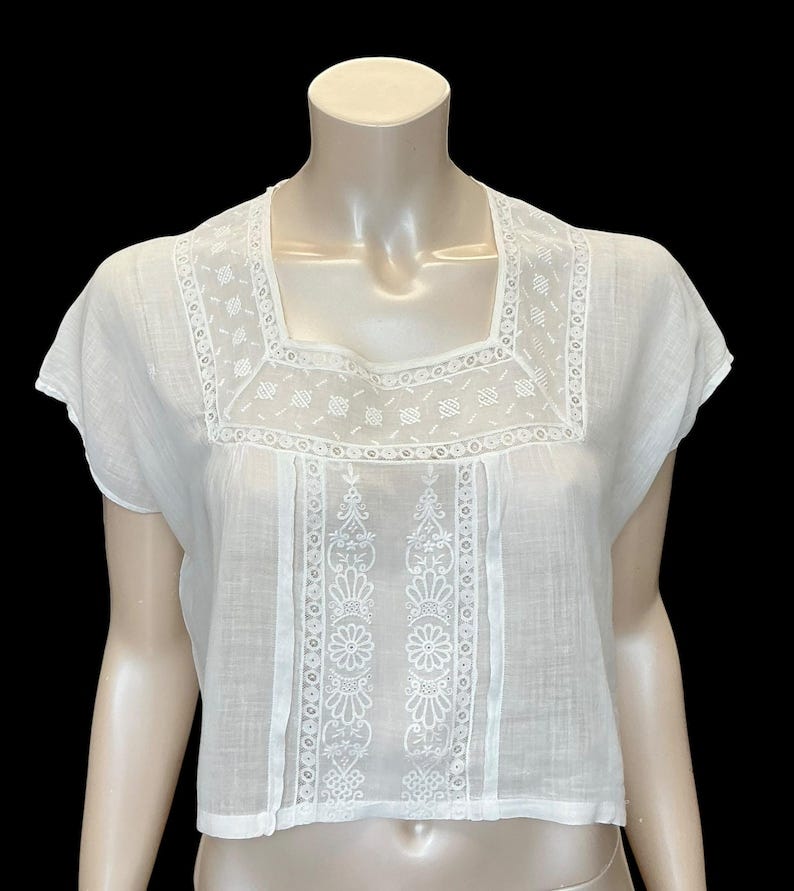 May include: A white short-sleeved blouse featuring a square neckline and intricate white embroidery. The garment displays a detailed floral pattern on the front panel and a geometric border around the neck, crafted from a lightweight fabric.