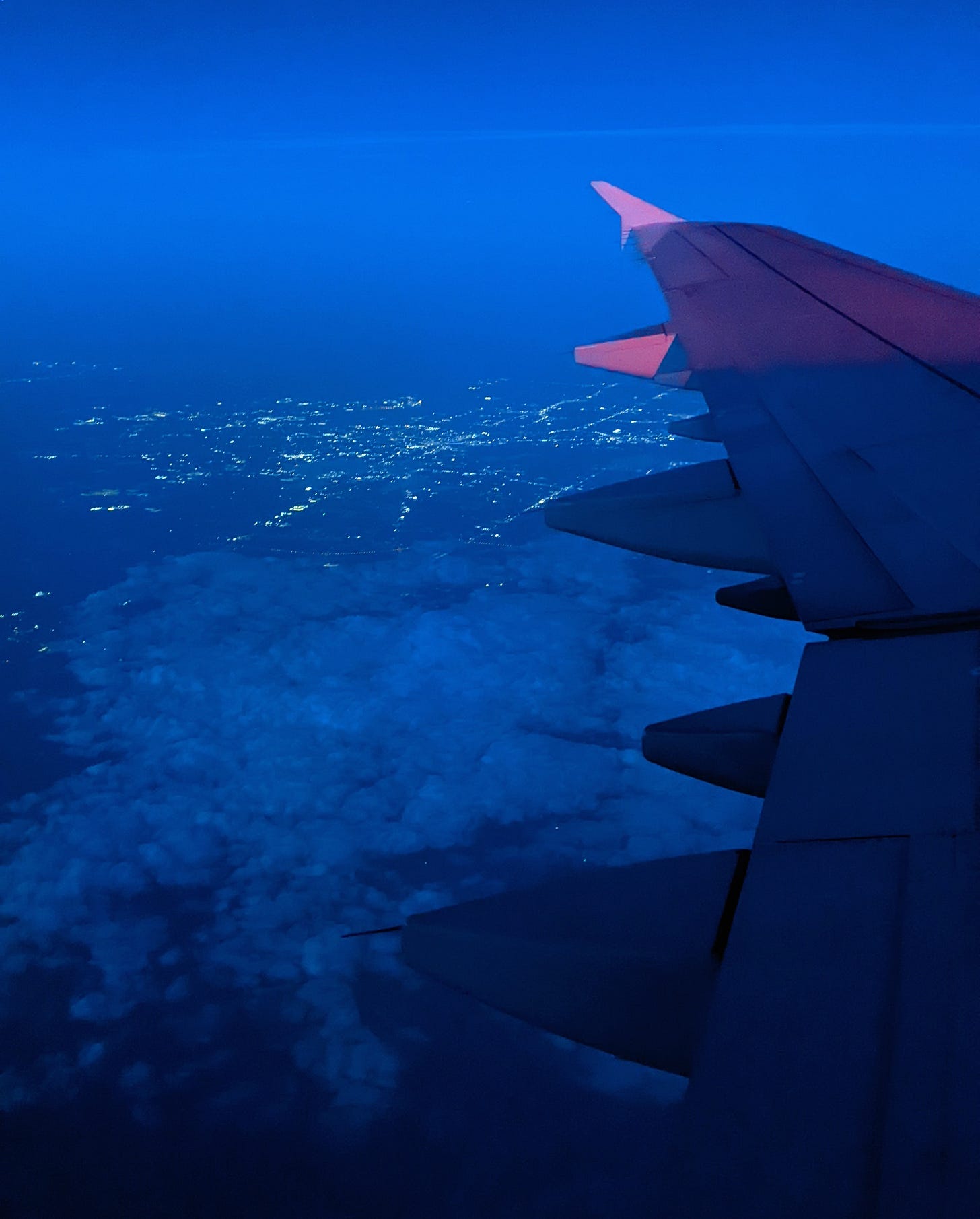 wing of a plane in flight above a city at night, with clouds covering the lower half