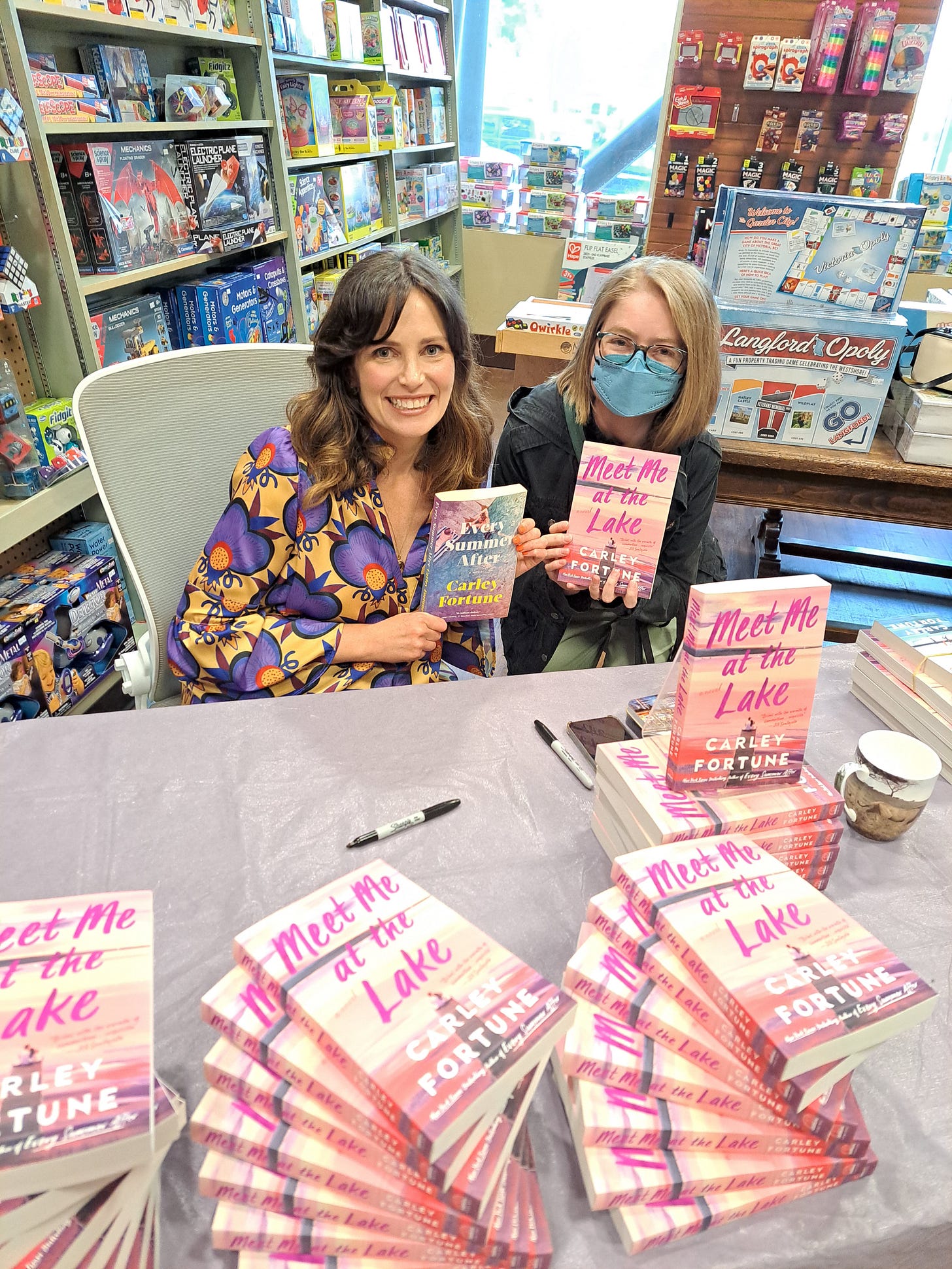 Author Carley Fortune sitting behind table holding a book and posing for a photo with me, a young blonde woman