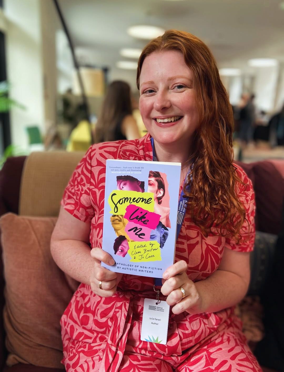 Julie, a woman with long red hair, blue eyes, and wearing a bright pink patterned jumpsuit, holds up Someone Like Me, which has a blue cover with pink and green blocks behind the title text. 