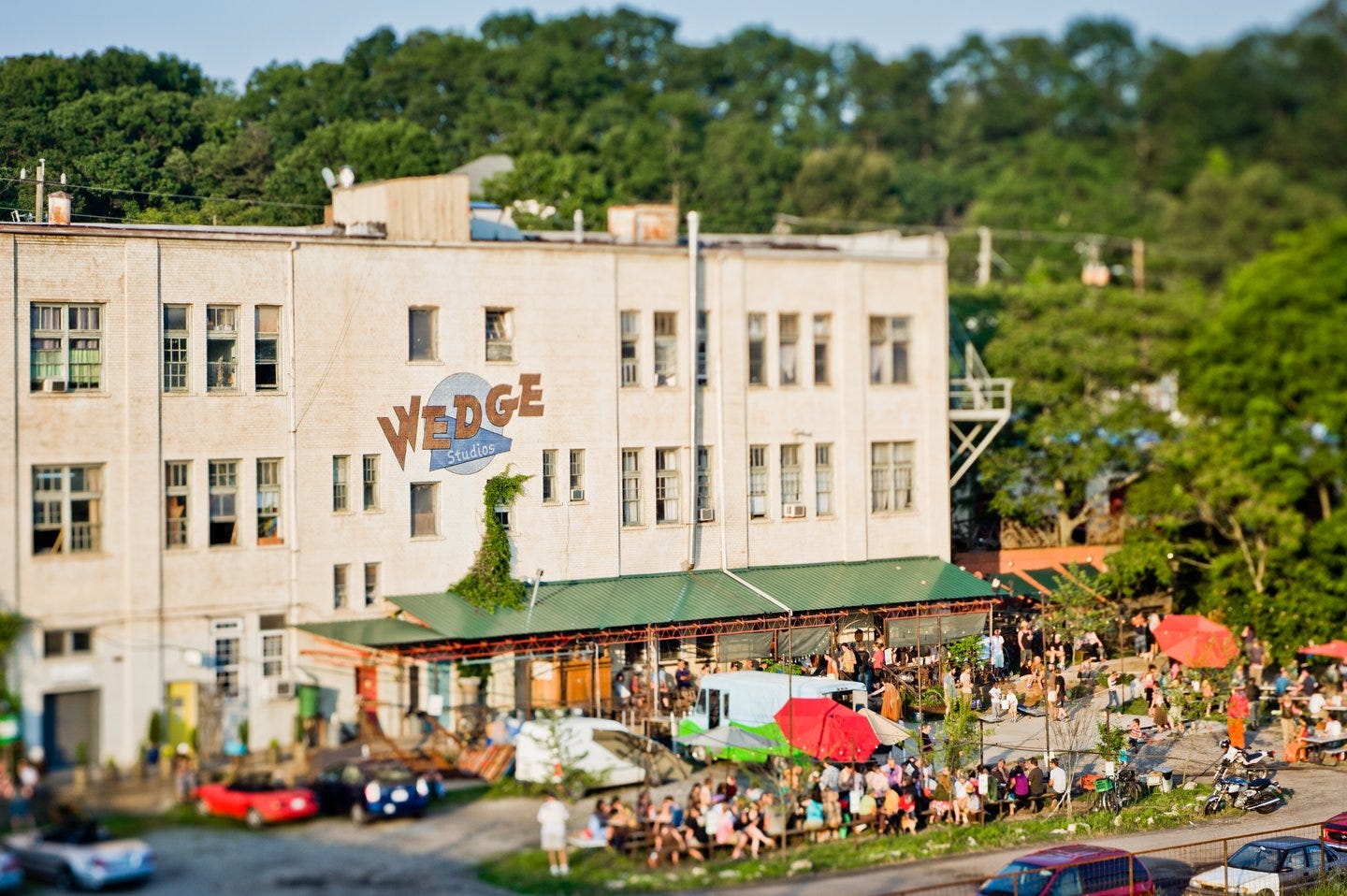 A lively outdoor scene at Wedge Studios, featuring a crowd enjoying food and drinks, with colorful umbrellas and greenery in the background.