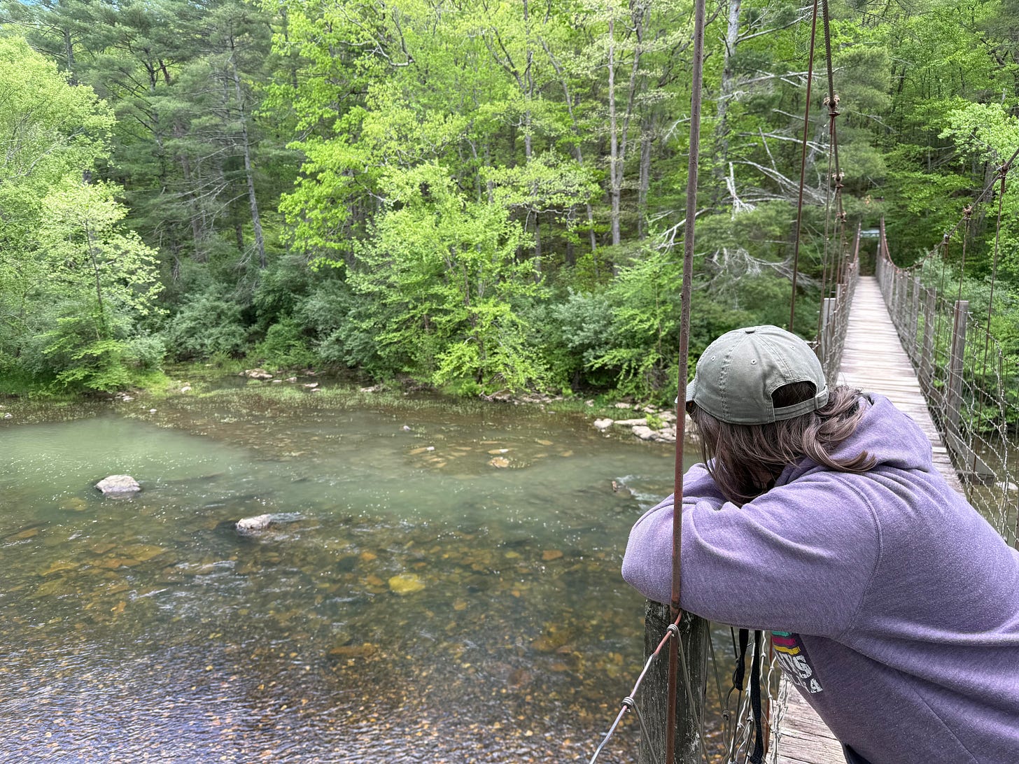 Woman in purple hoodie leaning on railing of a suspension bridge over the Maury River, surrounded by trees and nature