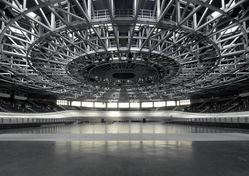 Imagem p&b da pista com um teto alto redondo com vigas metálicas expostas. Ao redor do palco, há uma pista de bicicleta oval com paredes altas e escuras.
