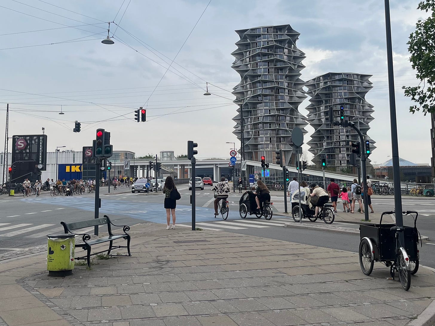 An urban intersection with two jagged-shaped highrise buildings in the background. Two cargo bikes and another bike are waiting to cross from the near side, and there are many bikes waiting to cross on the far side. There is a wide, blue painted area on the ground to indicate the bike lanes, and there is a parked cargo bike in the foregrond. To the left, in the background, are "S" signs indicating the presence of an S-train station, and an IKEA store, indicated by large "IKEA" in yellow on blue background