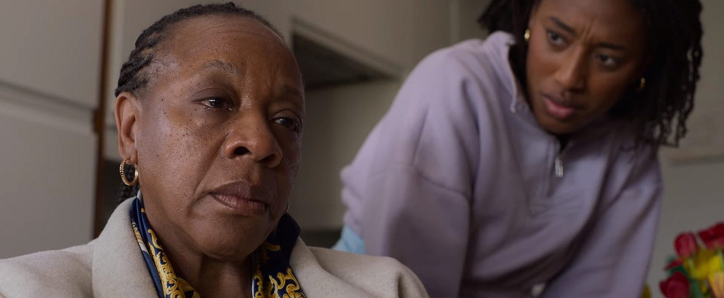 Still from the 2024 film Hard Truths. The image features two women in an emotional and tense moment. The older woman in the foreground wears a cream-colored coat, a patterned scarf, and gold hoop earrings. Her expression is somber and reflective. In the background, a younger woman dressed in a lavender hoodie leans forward, gazing at her with a concerned and inquisitive expression. The setting is an indoor space, with subtle details like a vase of flowers partially visible on the right.