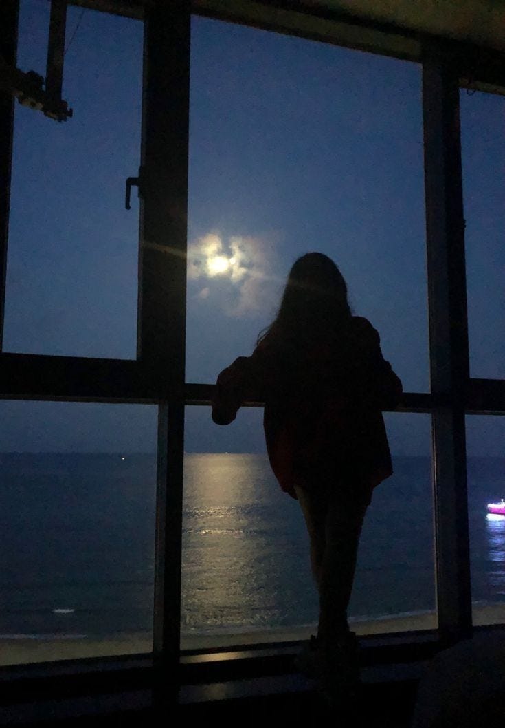 This may contain: a woman standing in front of a window looking out at the ocean under a full moon