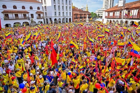 A large crowd of people many of them waving Colombian flags and dressed in its red, blue and yellow colours pack into a square