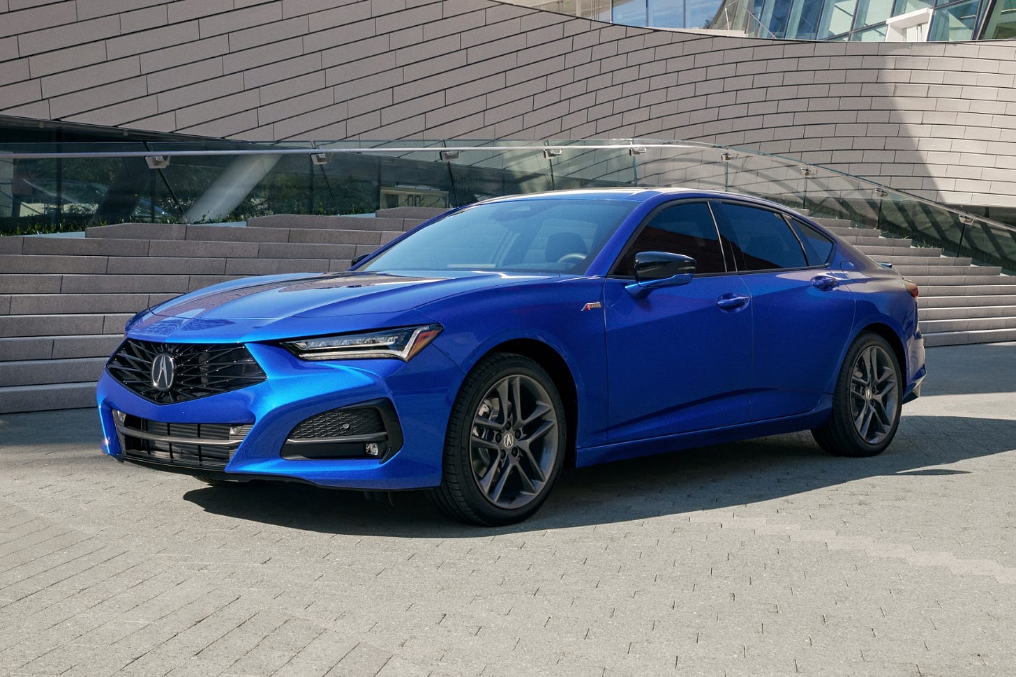 A blue 2024 Acura TLX A-Spec parked on gray pavers in front of a building with dramatic curved architecture.