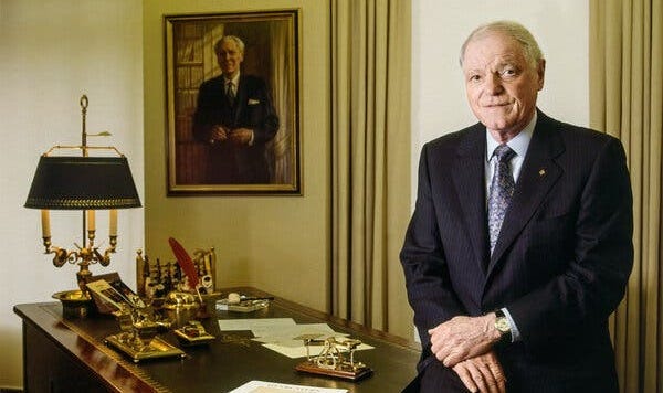 A man, staring into the camera, wearing a dark pinstriped suit and standing next to a wooden desk