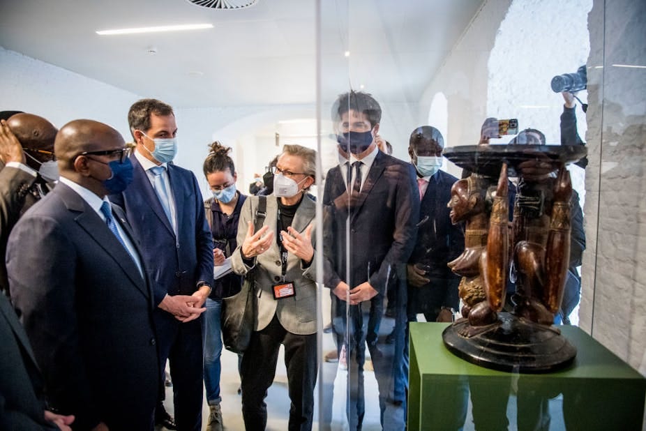 Men and women wearing face masks stand in front of a wooden sculpture of two figures, one bare-breasted, with concave trays at the top and bottom. It is inside a glass display case Men and women wearing face masks stand in front of a wooden sculpture of two figures, one bare-breasted, with concave trays at the top and bottom. It is inside a glass display case