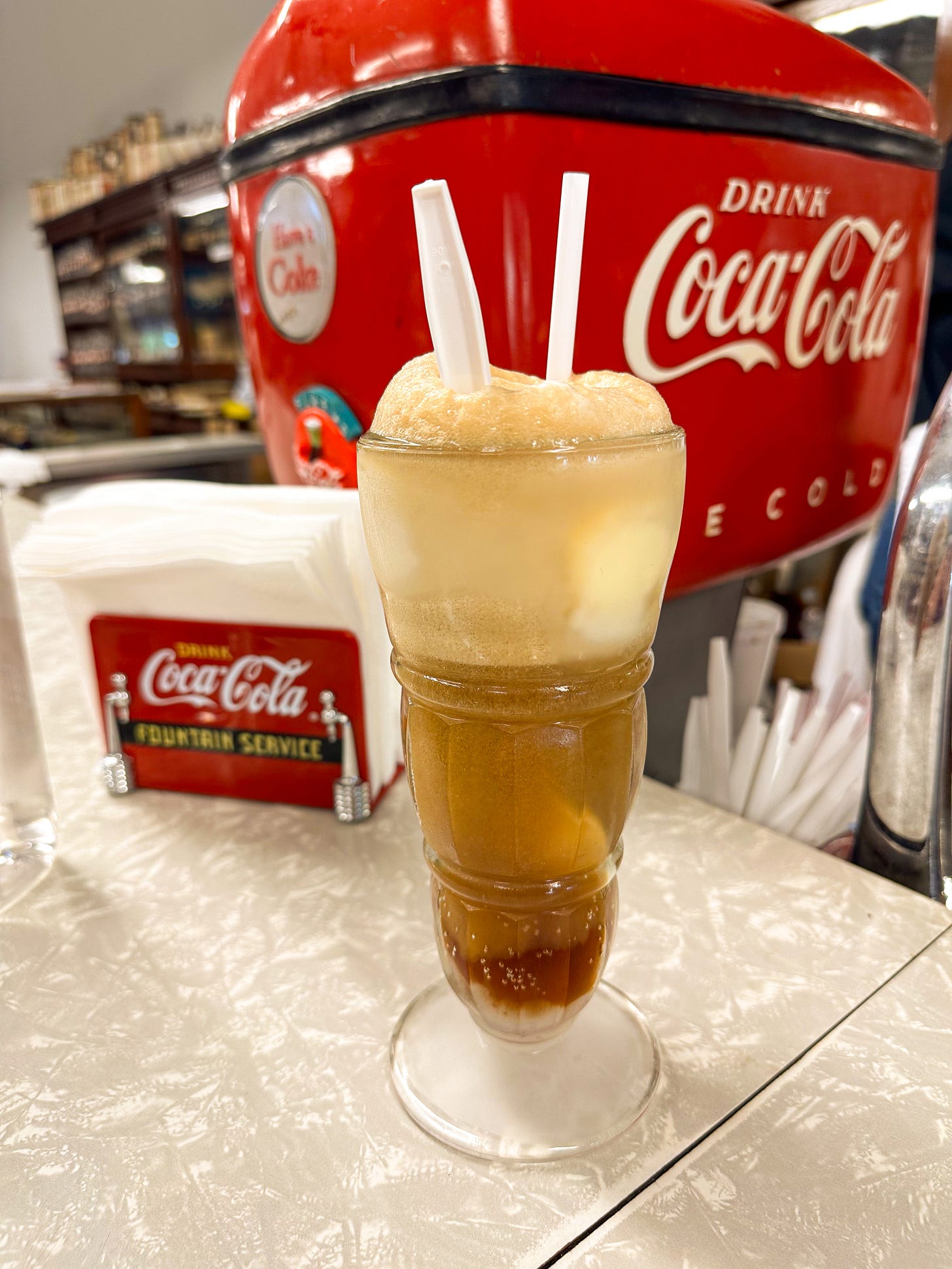 A coke float in a fancy glass, in front of an old Coke fountain