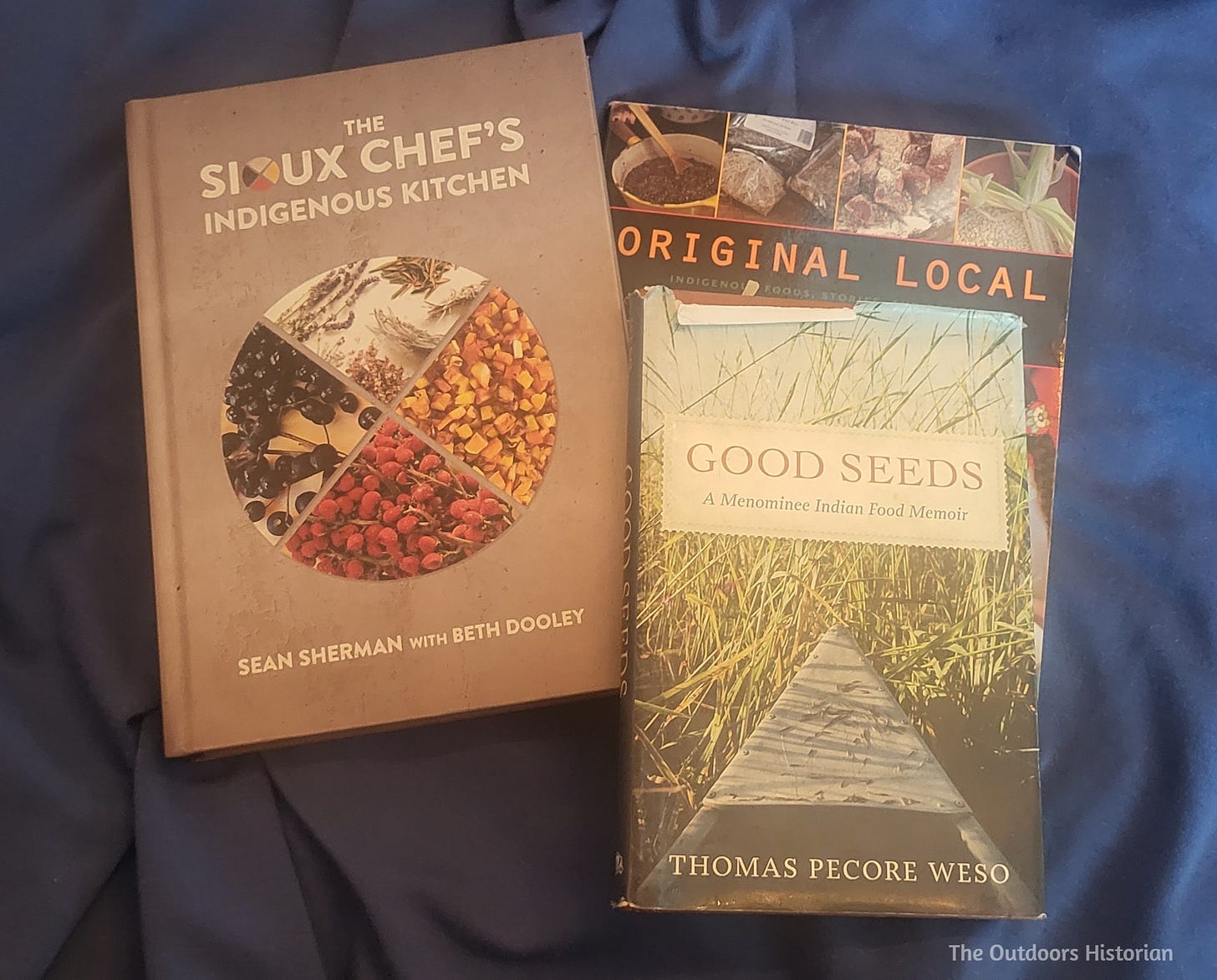 Three cookbooks by Sean Sherman, Thomas Pecore Weso, and Heid Erdrich are shown on a blue blanket.