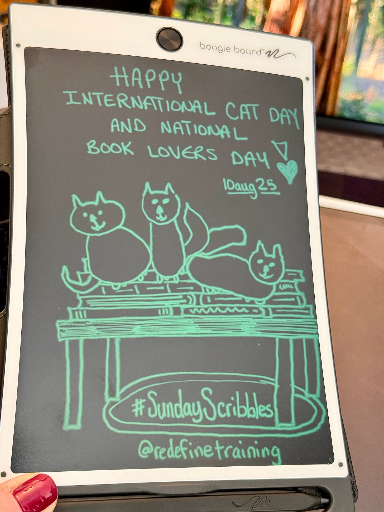 A digital sketch by Georgina Dean on a white Boogie Board. The sketch, drawn in a greenish-blue line on a dark screen, shows three cartoon cats sitting on a stack of books. The text reads "Happy International Cat Day and National Book Lovers Day, 10aug25," and "#SundayScribbles."