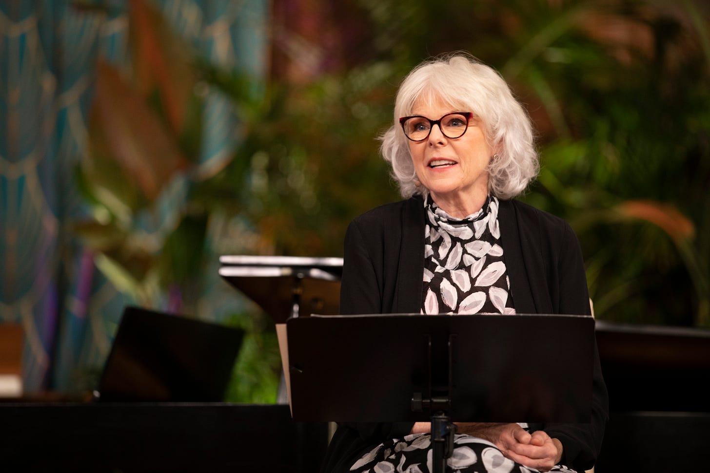 Photo of a woman with white hair and dark glasses speaking while seated at a podium. 