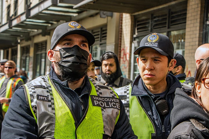 First image: A woman blows a cloud of vapour in the faces of police. A friend who was down there tells me her group was trolling the police the whole time, including taking drags from their vape and blowing it at the police. Second image: A person with a white shirt and a white wrap around their head and face and a black cap holds up a sign that reads: "Cops are better at committing crimes than solving them." Third image: An Indigenous person, back facing the line of police, is holding a bowl of smoking sage. After holding a cap overtop of the sage smoke, they turned the cap over, and smoke can be seen trickling upwards, out of the hat. Fourth image: A couple of police officers are seen at the front line. One is wearing a mask and looking dead ahead. The other is looking at something to the left of the camera's perspective.