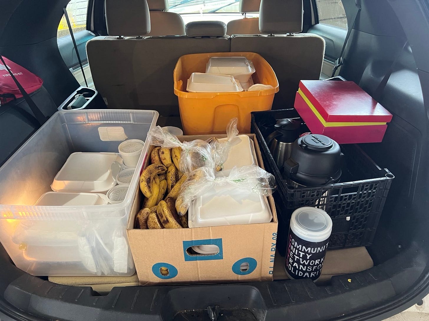 The trunk of a car with a clear plastic bin of white to-go boxes, an orange bin of white to-go boxes, a cardboard box of bananas and white to-go boxes, and a black crate with 3 drink carafes and a pink box of tea options