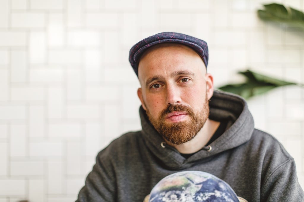 Author Justin Barker wearing grey sweatshirt and hat in front of a white tile background.