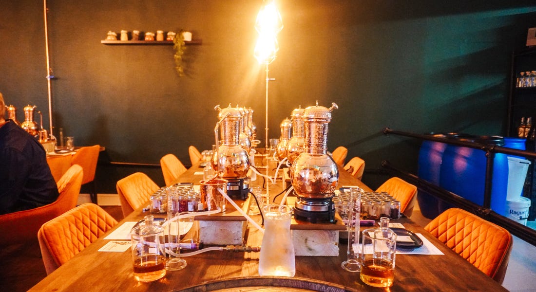 A wooden table with copper stills and rum making equipment on it, with orange chairs tucked into it A wooden table with copper stills and rum making equipment on it, with orange chairs tucked into it