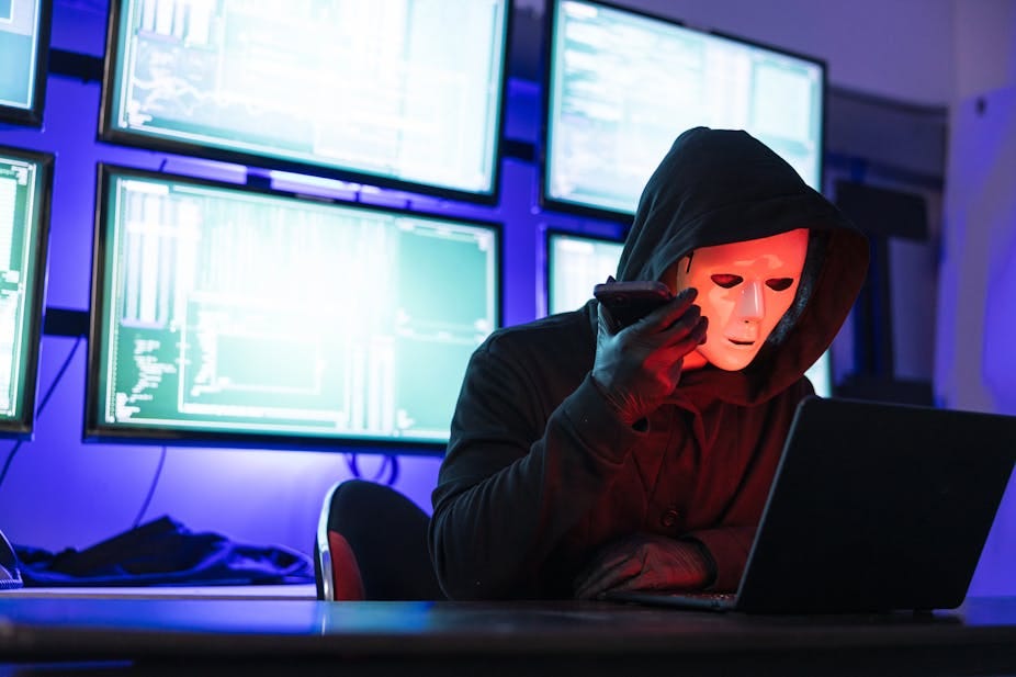 Hooded and masked person sitting at a computer, with digital screens behind on the wall