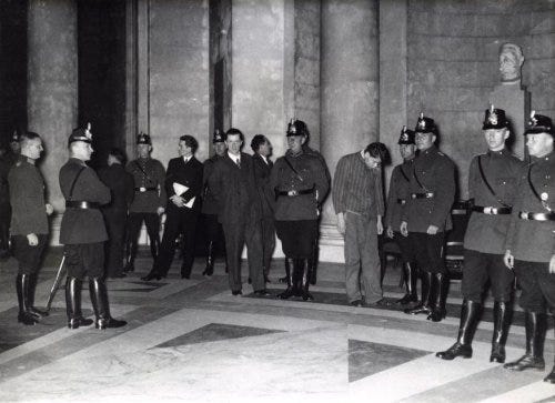 A black and white photograph with a dozen German soldiers and other men in different uniforms (police?). One man in the middle stands out: Marinus van der Lubbe, as the only one not in uniform, head bowed. Although not handcuffed, he looks defeated.