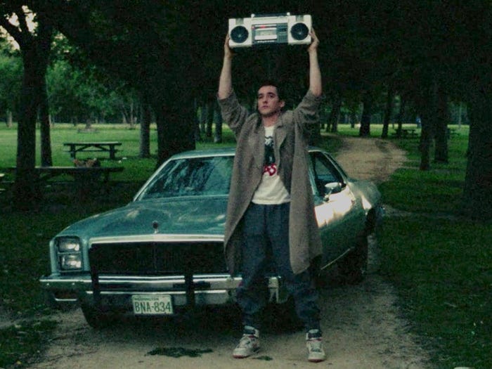 John Cusack in Say Anything holding a boombox above his head in front of a car.