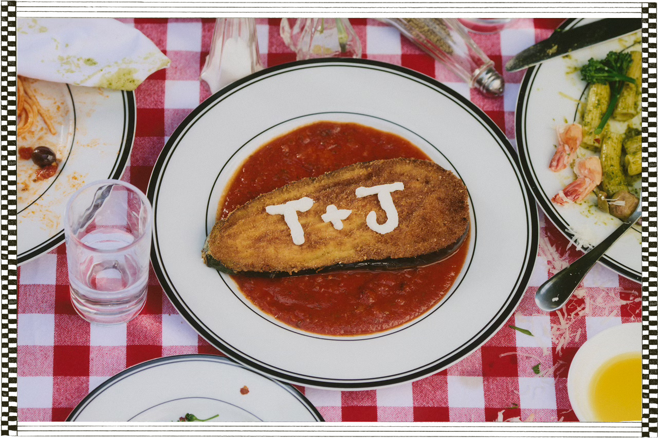 A breaded slice of eggplant rests in tomato sauce on a white plate, with “T + J” written in white sauce. The table is set with half-finished Italian dishes over a red-checkered tablecloth.