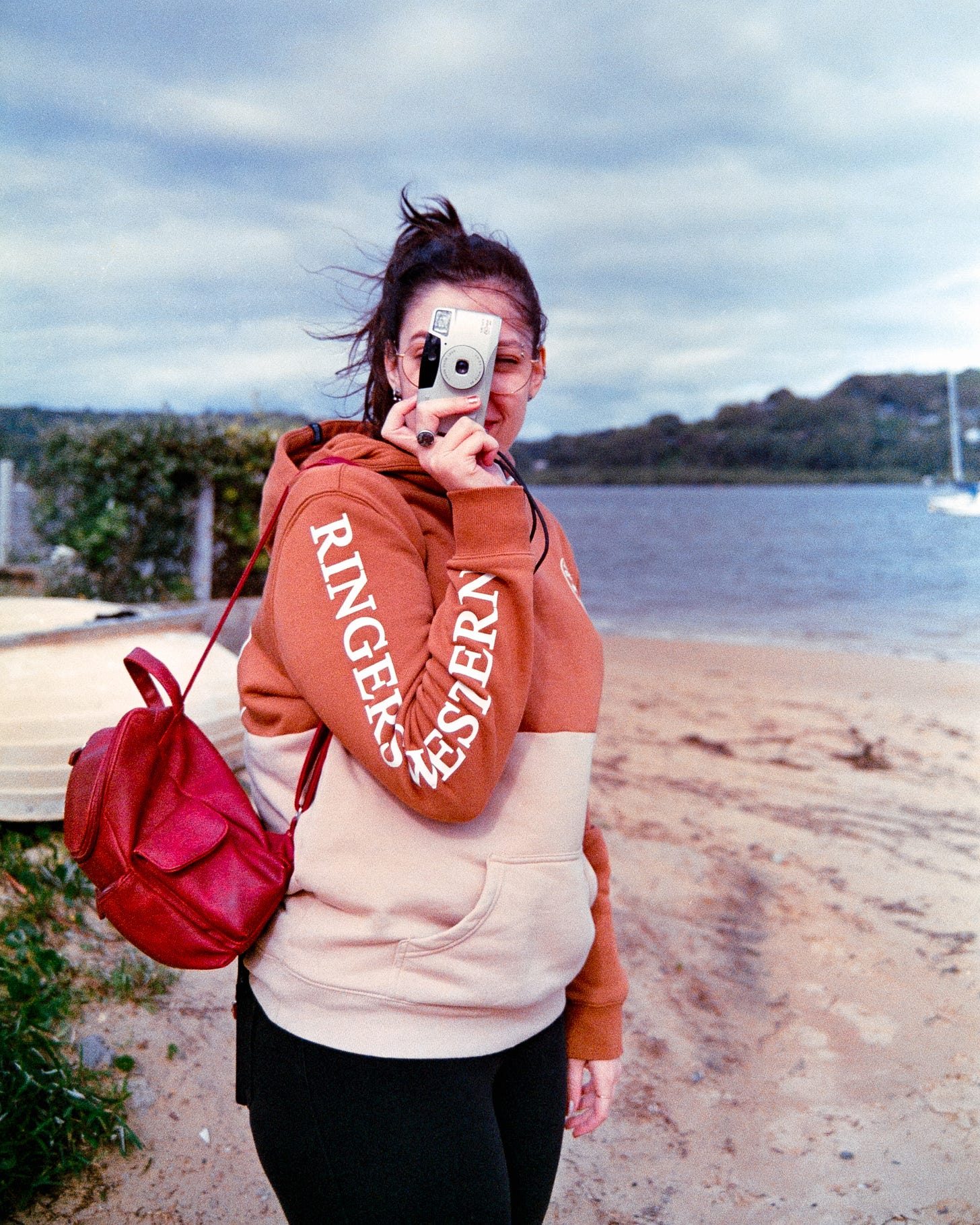 A photo of Josie smiling brightly with a thrifted film camera raised to her eye to take a photo. The wind is blowing her ponytail about, and her bright red bag stands out on the left side of the frame. A photo of Josie smiling brightly with a thrifted film camera raised to her eye to take a photo. The wind is blowing her ponytail about, and her bright red bag stands out on the left side of the frame.