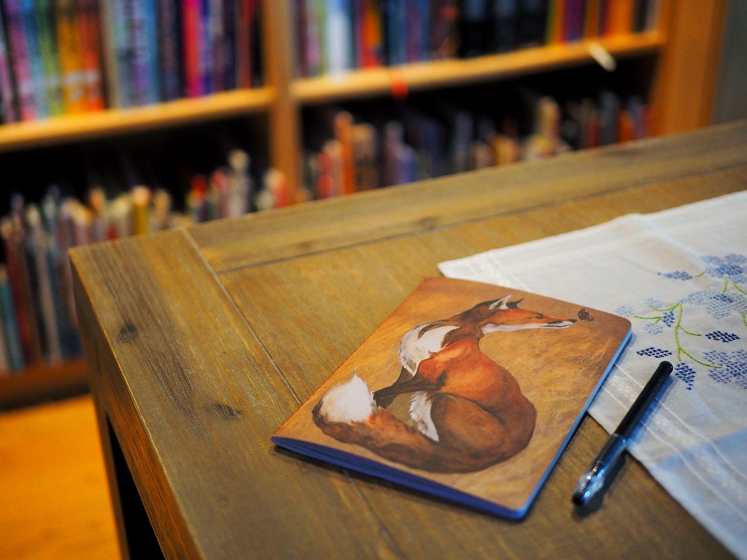 A photo of a thin journal with a gorgeous painting of a fox on it by the artist Jackie Morris. It sits on a table with a pen and there is an embroidered table runner. In the back ground, there is an out of focus bookcase. 