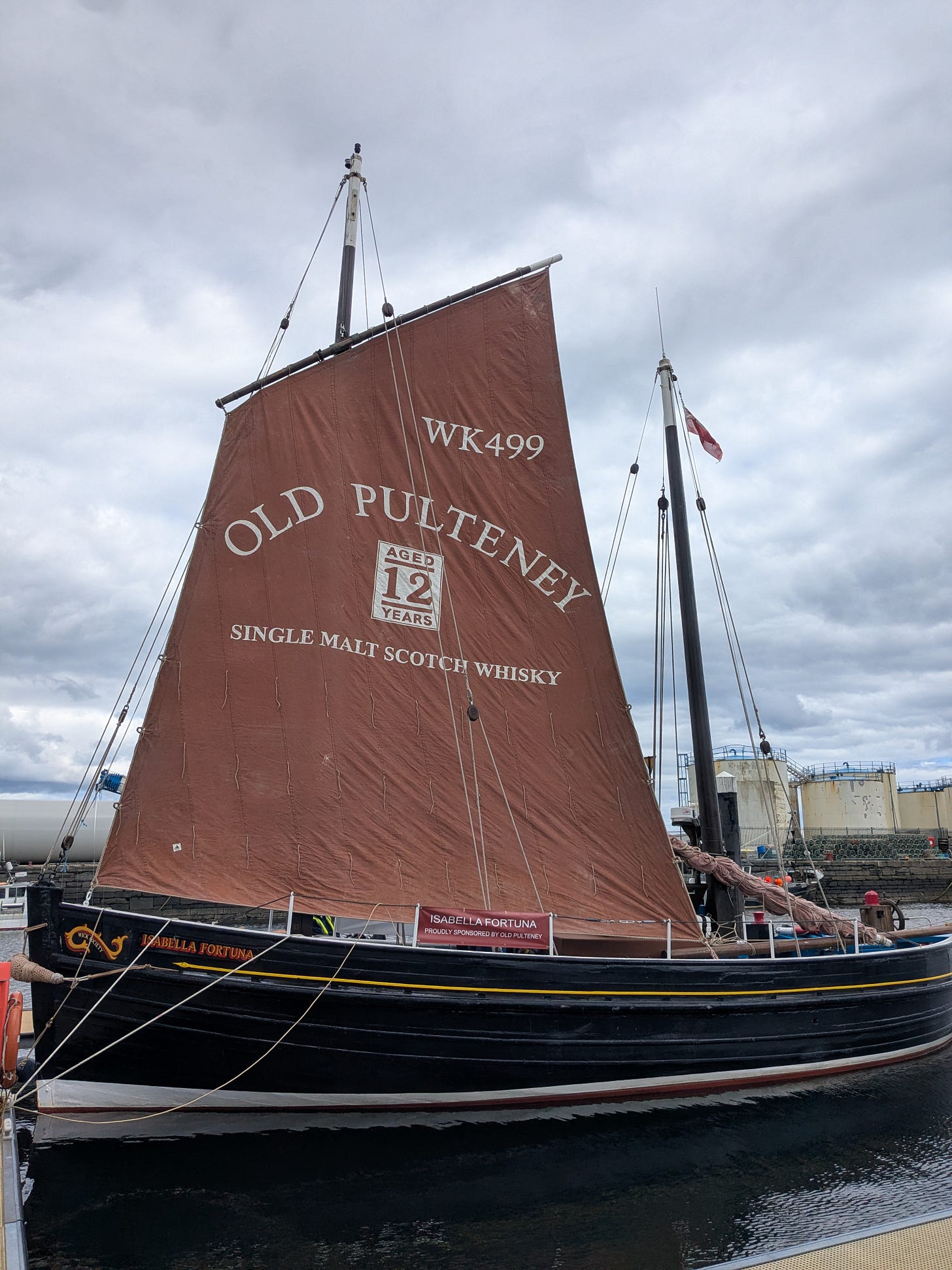 A detailed photograph of the traditional wooden Zulu fishing boat named Isabella Fortuna, moored in a harbour with stone walls and industrial elements in the background. The boat has a large, unfurled brown canvas sail that displays extensive white painted text, including 'WK499' and 'OLD PULTENEY 12 YEARS SINGLE MALT SCOTCH WHISKY'. The boat's name 'ISABELLA FORTUNA' is also visible in gold and red paint on the black hull. The image is taken from the side, showcasing the complex rigging and traditional construction.