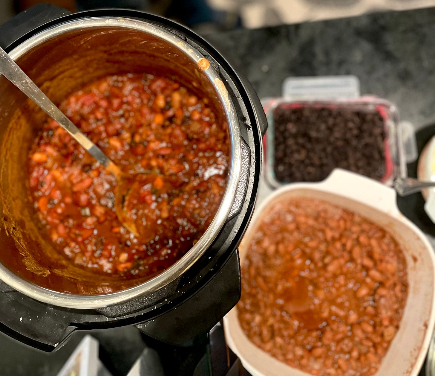 top left, an instant pot full of red bean-based chili, with a long-handled silver spoon sitting inside. Top right, a smaller rectangle container of black beans. Bottom right, a white square container of the author's finished pinto beans. Three bean dishes at a potluck.