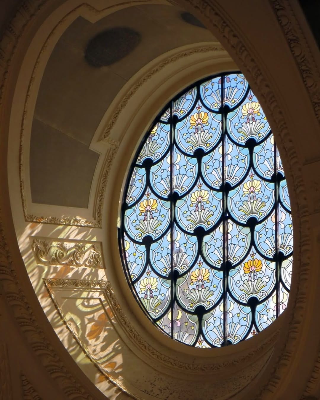 An ornate stained glass window at the Petit Palais, Paris. The window features a grid of blue, yellow, and white glass panels with floral and shell-like designs. The window is set within an arched, decorative stone frame with intricate molding. An ornate stained glass window at the Petit Palais, Paris. The window features a grid of blue, yellow, and white glass panels with floral and shell-like designs. The window is set within an arched, decorative stone frame with intricate molding.