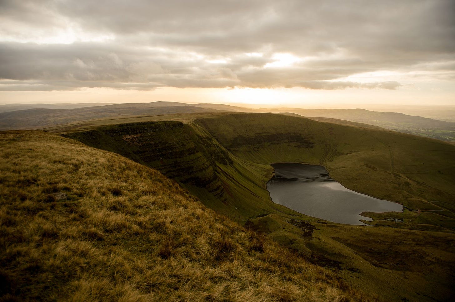 A sweeping view from a high, grassy ridge looking down into a deep basin holding a dark, still lake. Steep, green and golden-brown escarpment cliffs curve around the water. In the background, the sun breaks through dramatic, moody clouds, casting a warm, golden light over the distant rolling hills and foreground grasses.