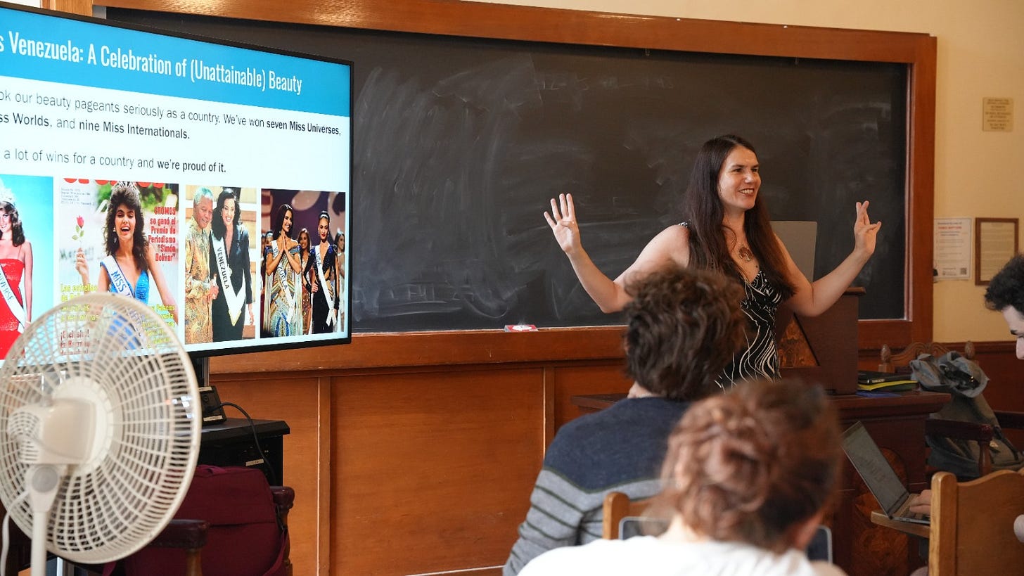 A woman stands in front of a blackboard and is smiling at the students seated in front of her. To her side there is a screen with photos of Venezuelan beauty queens across the decades.