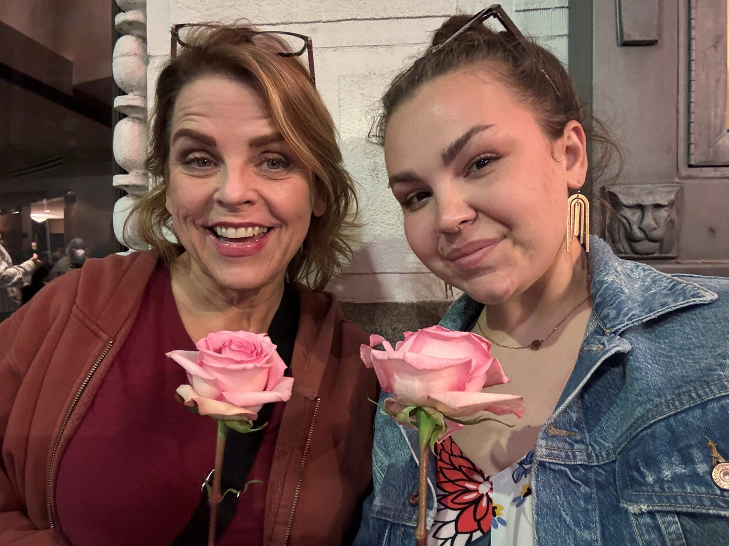 Two women, Julie Peters and her daughter, stand with roses in hand, posing in front of a building.